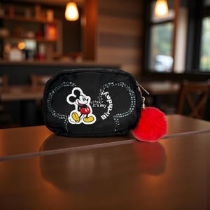 May include: Black zippered pouch with a Mickey Mouse design. The pouch features a Mickey Mouse graphic, the text "It's my Birthday!", and a red fluffy pom-pom attached to the zipper. The pouch is sitting on a brown surface.