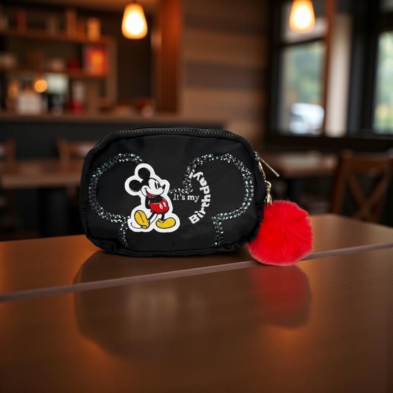 May include: Black zippered pouch with a Mickey Mouse design. The pouch features a Mickey Mouse graphic, the text "It's my Birthday!", and a red fluffy pom-pom attached to the zipper. The pouch is sitting on a brown surface.