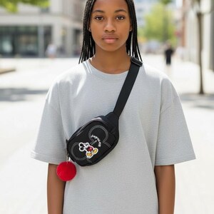 May include: A black fanny pack with a Mickey Mouse design and a red pom-pom. The bag is worn across the body and has a black strap. The person is wearing a gray t-shirt.