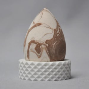May include: A teardrop-shaped makeup sponge with a marbled pattern in shades of brown and cream, resting on a white, geometric-patterned base. The sponge is designed for applying cosmetics.