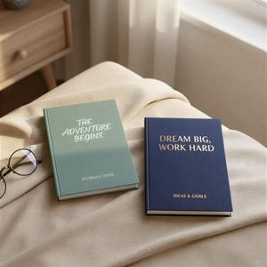 May include: Two hardback journals with motivational quotes. One is sage green with "The Adventure Begins" and "Journal 2026" on the cover. The other is navy blue with "Dream Big, Work Hard" and "Ideas & Goals".