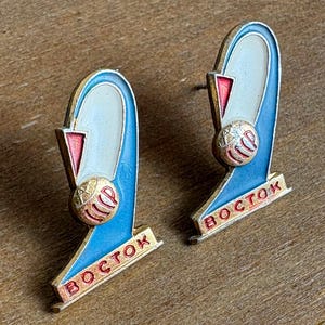 Puede incluir: Par de pines Vostok vintage. Cada pin presenta un diseño arqueado azul y blanco con un acento rojo y una esfera dorada. La palabra "BOCTOK" está escrita en rojo sobre una base dorada.