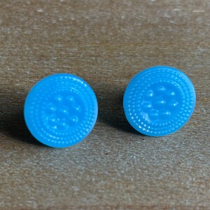 May include: Two light blue, round stud earrings. Each earring has a textured, dotted outer ring and a central design of raised dots. The earrings are displayed on a wooden surface.