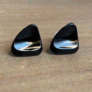 May include: A pair of black, triangular-shaped stud earrings. Each earring has a glossy finish with a gold-colored accent. The earrings are displayed on a light brown wooden surface.