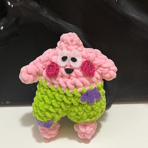 以下が含まれることがあります： 手作りの編みぐるみキャラクター、漫画のヒトデのフィギュア。フィギュアはピンク色で、緑色のショートパンツ、赤い頬、紫色のアクセントが付いています。フィギュアは笑顔で、柔らかい糸で作られています。