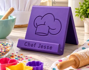 Op de afbeelding: Een paarse A-vormige tablet- of kookboekstandaard met een in reliëf aangebrachte koksmutspictogram op het voorpaneel en de tekst "Chef Jesse" gegraveerd op de onderste rand, gemaakt van een glad materiaal met een matte afwerking.