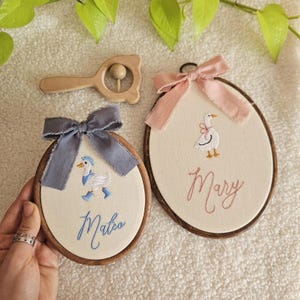 Könnte beinhalten: Zwei bestickte Kinderzimmer-Reifenkunstwerke mit Holzrahmen, die ein Gänsemotiv und die Namen "Mateo" und "Mary" zeigen. Eine Holzrassel und Schleifen in Rosa und Blau vervollständigen die Baby-Themen-Dekoration.