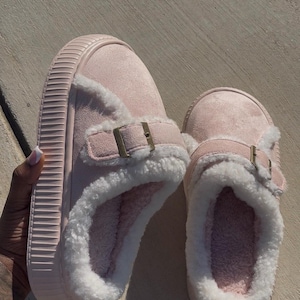 May include: A pair of light pink platform slippers with a faux suede exterior and a fluffy white interior lining. Each slipper features a strap with a gold-colored buckle. The soles have a ridged design.
