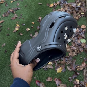 May include: Black clogs with star and chain embellishments. The clogs have a sporty design with the word "SPORT" printed on the side. The clogs are held by a person.
