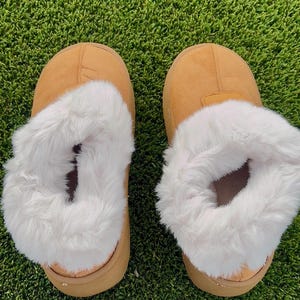 May include: A pair of tan-colored slippers with a fluffy white interior. The slippers are made of a soft material and are designed for indoor use. The slippers are placed on a green surface.