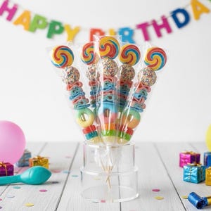 May include: A bouquet of five candy skewers individually wrapped in clear plastic. Each skewer features a colorful spiral lollipop, a round sprinkle-covered candy, blue and pink ribbon-shaped gummies, and a ring-shaped gummy candy at the base.