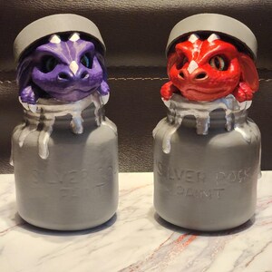 May include: Two decorative jars with dragon heads. One dragon is purple, the other is red. Both are emerging from gray jars with the text "SILVER ROCK PAINT". The jars have silver paint dripping down the sides.