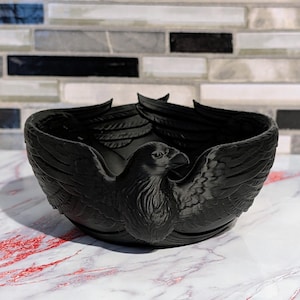 May include: A matte black bowl featuring a sculpted raven design integrated into the exterior walls. The bird's wings extend along the sides of the bowl, while the head and chest are prominently displayed on the front, creating a textured surface.