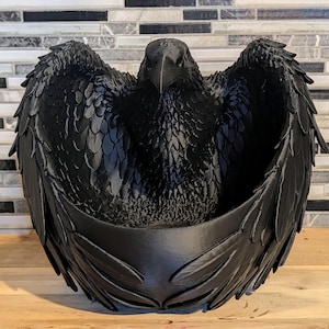 May include: A matte black raven-shaped bowl featuring detailed sculpted feathers across the wings and chest. The bird is depicted with its wings partially spread, creating a deep, rounded basin in the center for storage or display purposes.