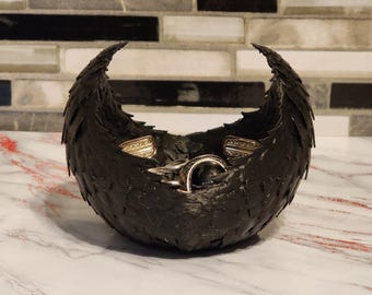May include: A black, decorative bowl with a textured, feather-like design. The bowl is filled with several rings. The bowl is sitting on a white and gray marble surface, with a black, gray, and white tile background.