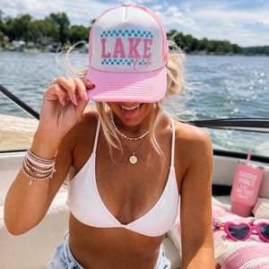 May include: A pink and white trucker hat with the words "LAKE COLA" in a blue and pink design. The hat is worn on a boat on a sunny day. The hat has a mesh back.