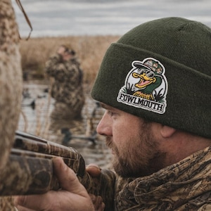 Może przedstawiać: Myśliwy w zielonej czapce z naszywką "Fowlmouth" przedstawiającą kreskówkową kaczkę w okularach przeciwsłonecznych. Myśliwy celuje z kamuflażowej strzelby w bagnistym otoczeniu.