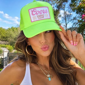 Puede incluir: Una gorra de estilo camionero en color verde neón con un parche rectangular en la parte frontal. El parche tiene fondo blanco, borde rosa y el texto Coors Banquet en letras rosas y marrones. La gorra cuenta con una malla en la parte trasera.