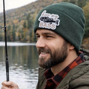 Może przedstawiać: Ciemnozielona czapka beanie z białą naszywką z czarnym basem i napisem "Kiss My Bass" w kursywie. Czapka wykonana jest z miękkiego materiału dzianinowego, idealna do wędkowania lub aktywności na świeżym powietrzu.