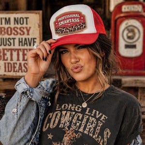 May include: A red and white trucker hat with the text "PROFESSIONAL SMARTASS" on the front. The hat features a leopard print design. The person is wearing a black t-shirt with the words "LONG LIVE COWGIRLS".