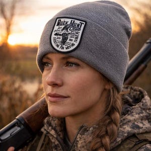 Może przedstawiać: Szara czapka z czarno-białą naszywką z napisem "Duck Hunt Club" z grafiką kaczki. Czapkę nosi osoba w kamuflażowej kurtce i trzymająca strzelbę.