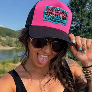 May include: A bright pink and black trucker hat featuring a rectangular patch with a leopard print background, a teal border, and the text Pretty Good AT BAD DECISIONS in pink and teal script and block lettering on the front panel.