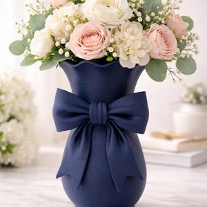 Könnte beinhalten: Eine marineblaue Vase mit einer großen Schleife enthält einen Strauß weißer, rosa und cremefarbener Blumen. Das Arrangement umfasst Rosen, Hortensien und Schleierkraut und schafft eine weiche, elegante Präsentation. Die Vase hat einen gewellten Rand.
