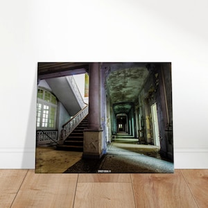 Könnte beinhalten: Leinwanddruck eines verlassenen Gebäudes. Das Bild zeigt einen langen Flur mit abblätternder Farbe, Säulen und einer Treppe. Die Farbpalette ist gedämpft mit Grautönen, Blau und Braun. Der Druck wird auf einem Holzboden präsentiert.