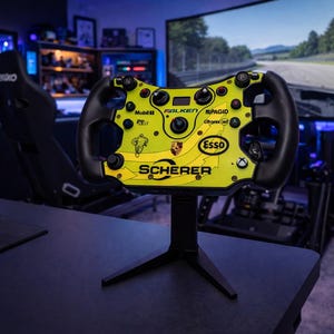 Può includere: Un volante da corsa giallo e nero con vari loghi di sponsor, tra cui Esso e Porsche, montato su un supporto nero. Il volante è in una configurazione di gioco con un sedile da corsa e un grande schermo che mostra una pista.