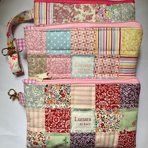 May include: Three patchwork zipper pouches in pink, red, and floral patterns. Each pouch features a different fabric design, including stripes, polka dots, and floral prints. The pouches have a pink zip and a wrist strap with a gold clasp. The label reads "Lunara by Lucy."