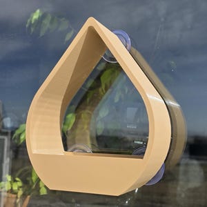 May include: A beige, teardrop-shaped plant holder attached to a window with suction cups. The holder has a flat base and a triangular top, creating an open space for plants. The window reflects the surrounding environment.
