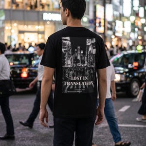 以下が含まれることがあります： 背面に長方形の白黒グラフィックプリントが施された黒の半袖Tシャツ。プリントには賑やかな街の通りの風景が描かれており、白い大文字で「LOST IN TRANSLATION」および「ANTHCRIPT DEPT」というテキストが印刷されています。