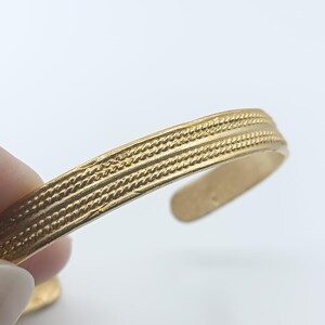 May include: Gold-coloured cuff bracelet with a textured, braided design. The open cuff design allows for easy wear. The bracelet is shown against a white background.