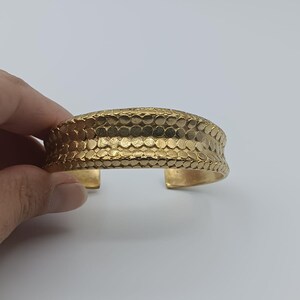 Puede incluir: Un brazalete dorado con un diseño texturizado y martillado. La banda ancha presenta un patrón repetitivo de pequeñas hendiduras circulares. El diseño abierto facilita su uso. El brazalete es una pieza llamativa.