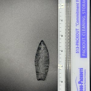 May include: A dark grey, hand-knapped arrowhead, approximately 9 cm long, lies on a grey surface. The arrowhead has a textured surface and a sharp point. A ruler is positioned next to the arrowhead for scale.