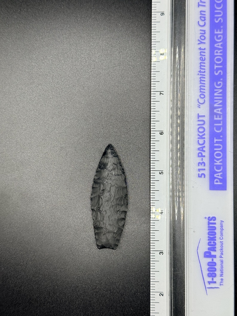May include: A dark grey, hand-knapped arrowhead, approximately 9 cm long, lies on a grey surface. The arrowhead has a textured surface and a sharp point. A ruler is positioned next to the arrowhead for scale.