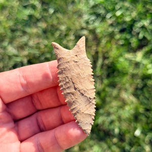 May include: A tan, bifacial stone arrowhead featuring a serrated edge and a bifurcated base. The surface displays a textured, knapped finish with visible flake scars across the entire body of the point, tapering to a sharp, pointed tip.