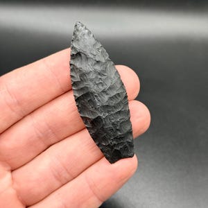 Puede incluir: Una punta de flecha negra, tallada a mano, con una superficie texturizada. La punta de flecha se sostiene en una mano sobre un fondo oscuro. La punta de flecha mide aproximadamente 10 cm de largo.