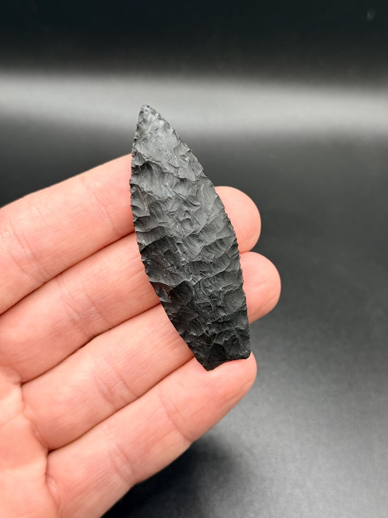 May include: A black, hand-knapped arrowhead with a textured surface. The arrowhead is held in a hand against a dark background. The arrowhead is approximately 10 cm long.