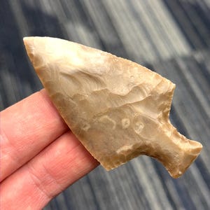 May include: A tan-colored, chipped stone arrowhead with a triangular shape and a notched base. The arrowhead has a textured surface and appears to be hand-crafted. The artifact is held in a person's hand.