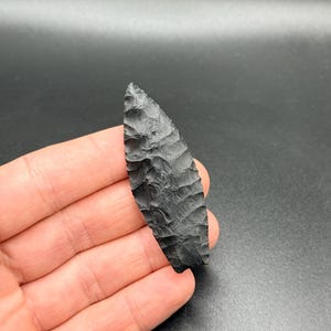 May include: A black obsidian arrowhead with a textured surface, held in a hand. The arrowhead is elongated with a pointed tip. The edges are finely worked, and the shape is symmetrical. The background is a dark, solid colour.