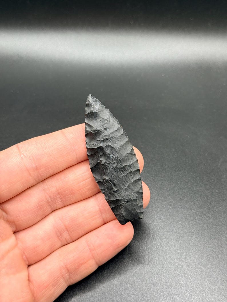 May include: A black obsidian arrowhead with a textured surface, held in a hand. The arrowhead is elongated with a pointed tip. The edges are finely worked, and the shape is symmetrical. The background is a dark, solid colour.