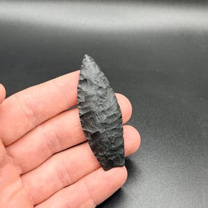 May include: A black, hand-carved arrowhead with a textured surface. The arrowhead is leaf-shaped, with a pointed tip and a wide base. The dark colour and intricate carving suggest a handcrafted item.