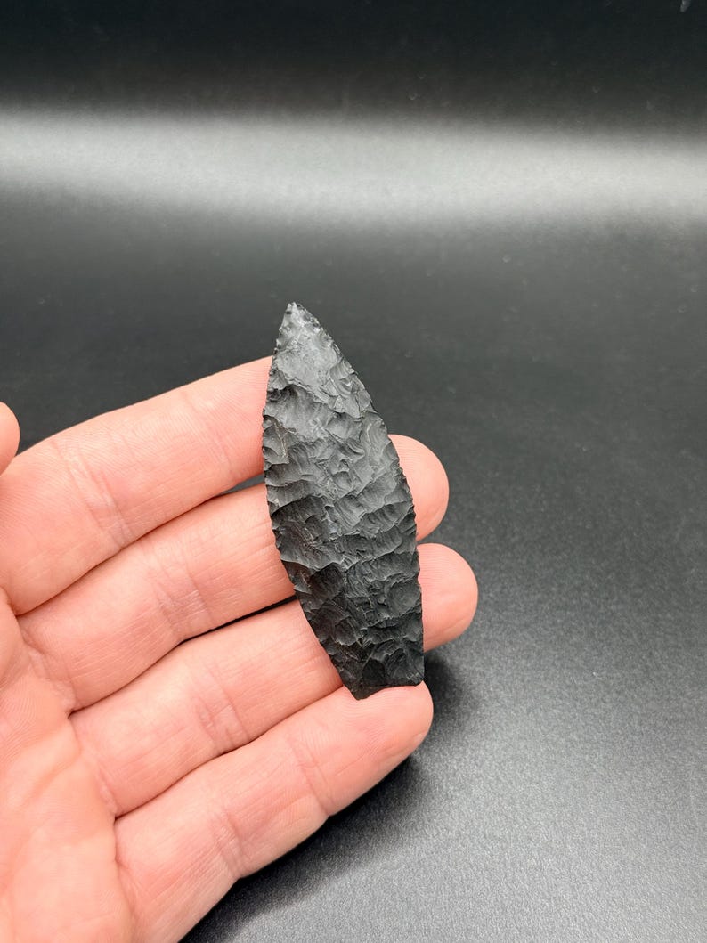 May include: A black, hand-carved arrowhead with a textured surface. The arrowhead is leaf-shaped, with a pointed tip and a wide base. The dark colour and intricate carving suggest a handcrafted item.