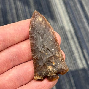 May include: A brown arrowhead with a textured surface and a sharp point. The arrowhead is held in a hand, showcasing its size and detail. The stone artifact has a chipped edge and a rough texture.