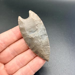 Puede incluir: Una punta de flecha gris con base dentada, sostenida en una mano. La herramienta de piedra tiene forma triangular con punta y muesca curva en la parte superior. La superficie muestra variaciones naturales de color y textura.