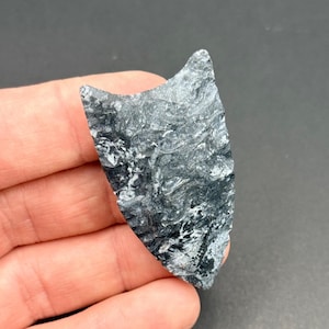 Puede incluir: Una pieza de piedra pulida con forma de escudo. La piedra presenta un patrón arremolinado de negro, gris y blanco, creando un efecto veteado. Los bordes son irregulares, lo que le da una apariencia natural y orgánica.