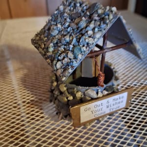 May include: A miniature wishing well featuring a roof and base covered in small, multi-colored pebbles. The structure includes a brown wooden support frame with a small bucket hanging from a string. A gold-colored sign reads: Go Out & Make Your Wishes Come True.