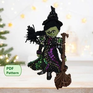 May include: A handmade witch doll with a black hat, dress, and broom. The witch has green skin, round glasses, and a black braided hairstyle. The dress is embellished with sequins in green and purple. A green label with the text "PDF Pattern" is in the lower left.