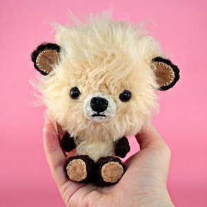 May include: A small, fluffy, cream-colored crocheted stuffed animal with black eyes and a black nose. The animal has brown ears with black trim and brown feet with brown stitching. The animal is being held in a person's hand.